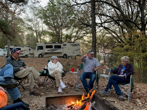 4 people sitting by a campfire with RVs in the background.
